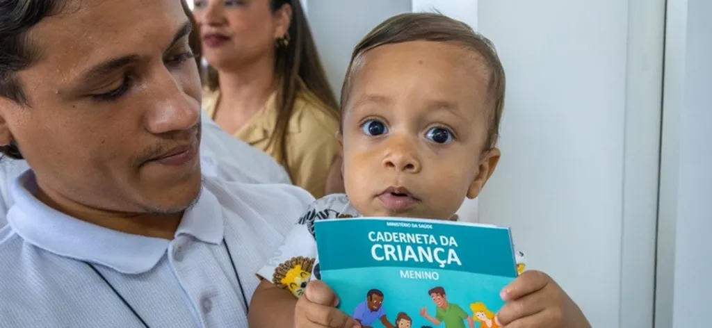Imagem mostra um homem com menino no colo, que segura uma caderneta da criança