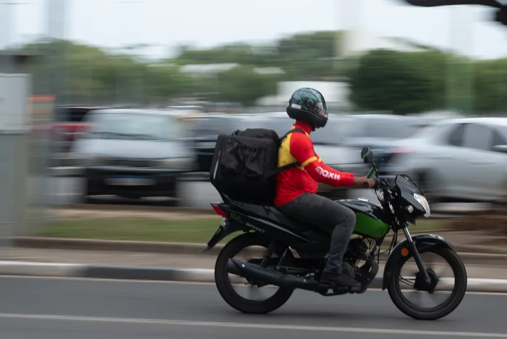 Imagem mostra motociclista pilotando. Ele veste camisa longa na cor vermelha e calça jeans escura