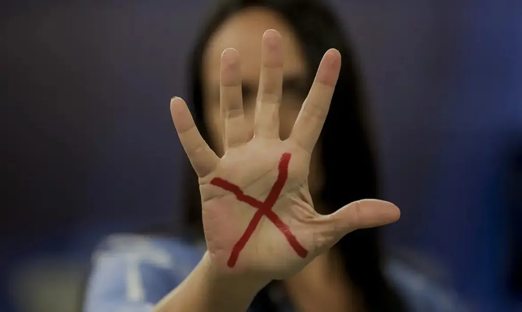 Imagem mostra mulher com a mão a frente do rosto, com um "X" em vermelho na palma da mão