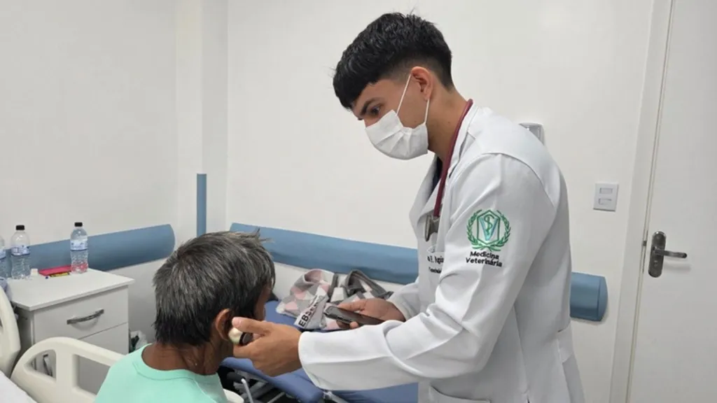 Imagem mostra acadêmico, de jaleco branco, aproximando caixinha de som ao ouvido de paciente indígena