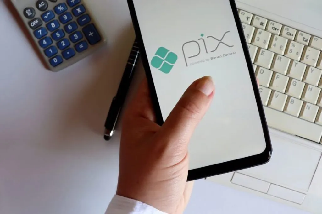 Imagem mostra mão segurando celular, com "Pix" aberto. Ao fundo, um teclado de computador, uma calculadora e uma caneta