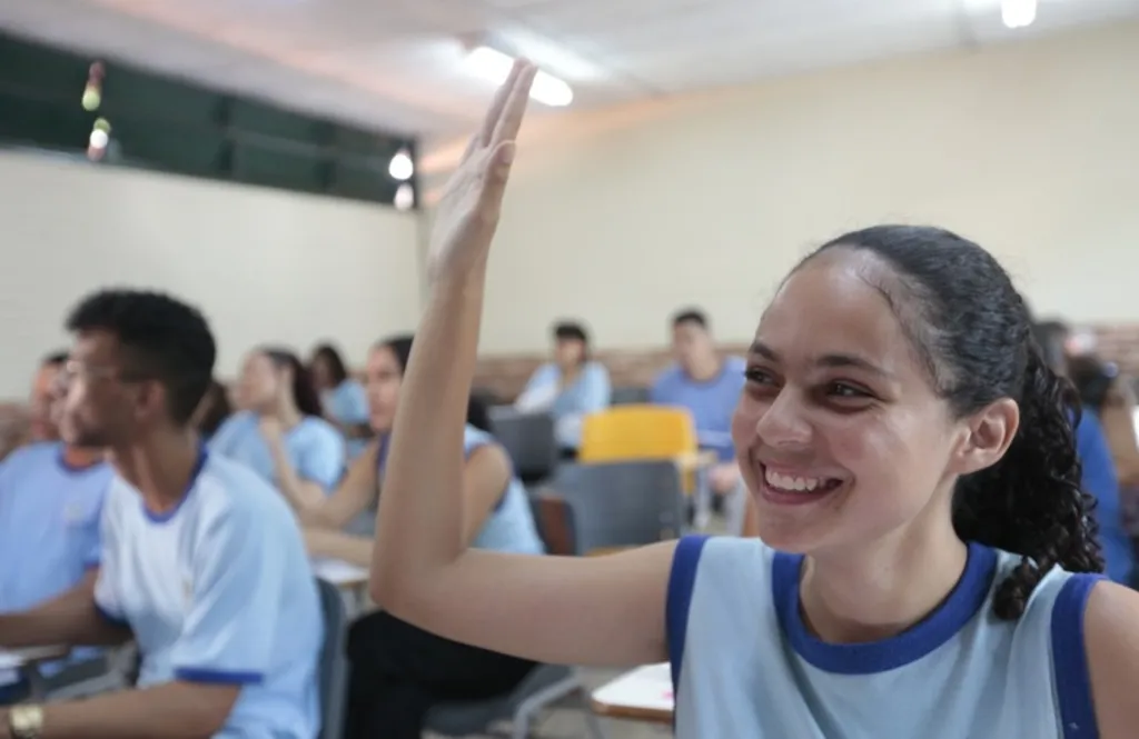 Imagem mostra alunos em sala de aula, com destaque para aluna sorrindo e com a mão direita levantada